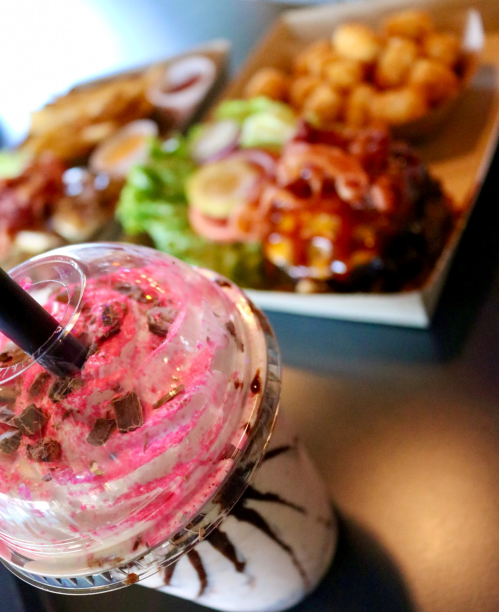 A colorful milkshake topped with whipped cream and chocolate pieces, with a plate of food in the background.