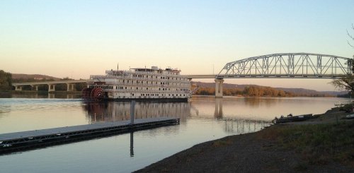 A riverboat with a paddle wheel docked near a bridge, reflecting on calm waters at sunset.