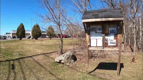 A wooden information kiosk with photos stands in a grassy area, surrounded by trees and a clear blue sky.