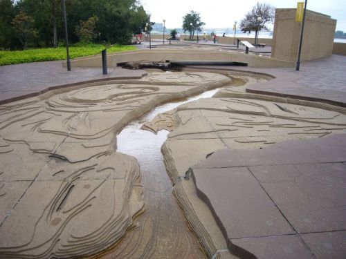 A sculptural water feature with flowing lines and a shallow stream, surrounded by greenery and pathways.