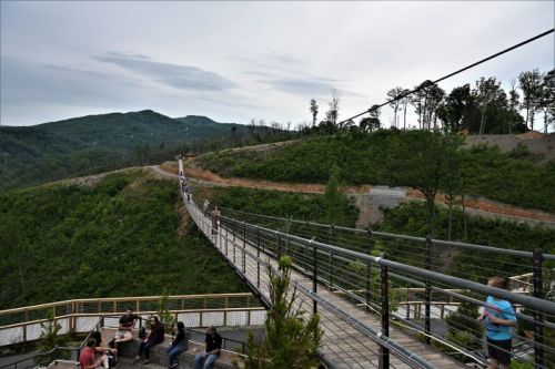 A scenic view of a suspension bridge with people, surrounded by green hills and a cloudy sky.