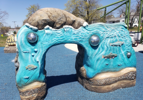 A colorful, textured rock archway with fish designs, set in a playground with blue rubber flooring.