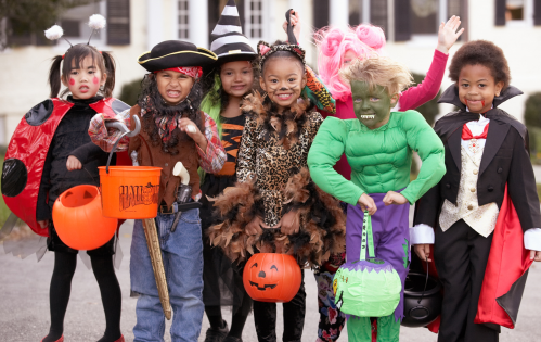 A group of children in colorful Halloween costumes, including a pirate, witch, and superhero, smiling together outdoors.