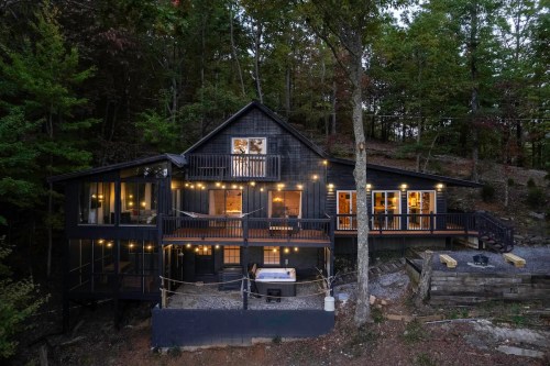 A modern black cabin surrounded by trees, featuring outdoor lighting and a hot tub on a gravel path.