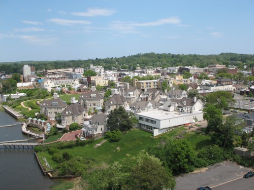 Aerial view of a coastal town featuring houses, greenery, and a waterfront with boats. Clear blue sky above.
