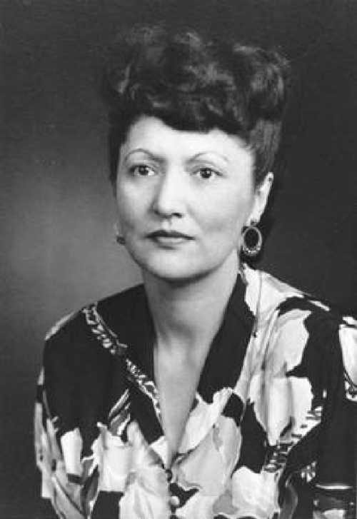 Black and white portrait of a woman with styled hair, wearing a floral blouse and earrings, looking directly at the camera.