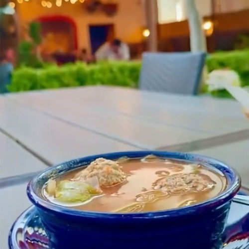 A bowl of soup with toppings, placed on a table in a cozy outdoor setting with greenery in the background.