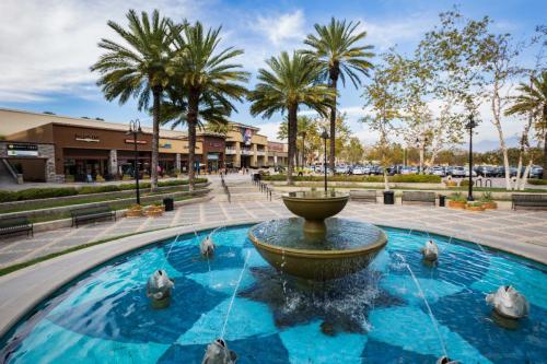 A fountain surrounded by palm trees and shops in a plaza on a sunny day. Benches line the walkway.