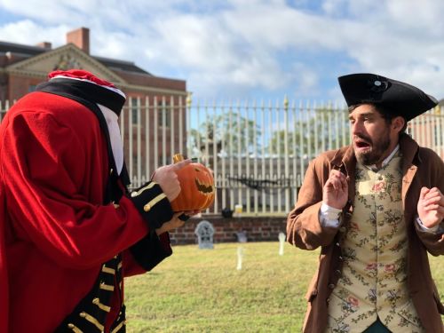 A headless figure in a red coat holds a carved pumpkin, while a man in a brown coat looks startled in a graveyard setting.