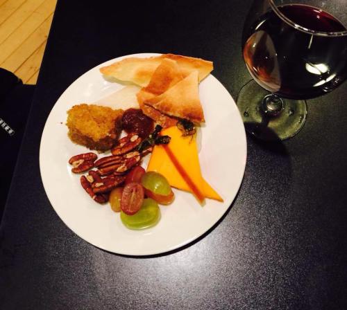 A plate with assorted snacks: crackers, cheese, nuts, grapes, and a glass of red wine on a black table.