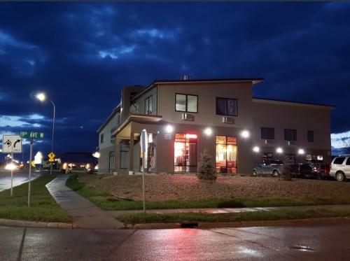 A modern building at dusk, illuminated by warm lights, with a cloudy sky and wet pavement reflecting the scene.