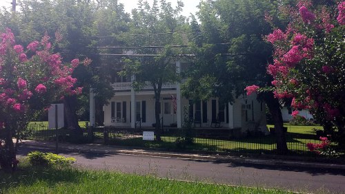A charming white house surrounded by vibrant pink flowers and lush greenery on a sunny day.