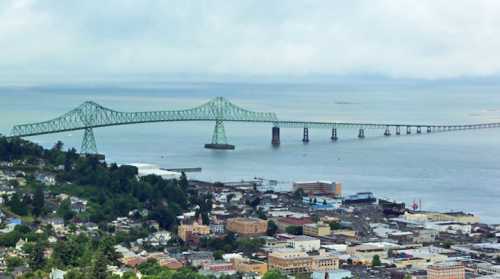 Aerial view of a green bridge spanning a river, with a coastal town and cloudy sky in the background.