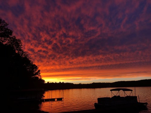 A vibrant sunset over a lake, with colorful clouds reflecting on the water and silhouettes of trees and boats.
