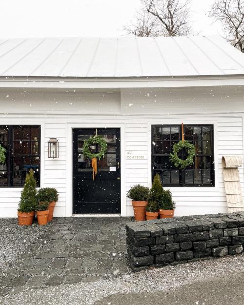 A charming white building with a black door, adorned with wreaths, surrounded by potted plants, in a snowy setting.