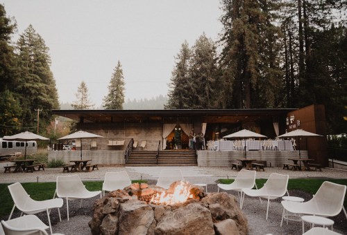 A cozy outdoor venue with a fire pit, surrounded by trees, featuring a modern building and seating areas.