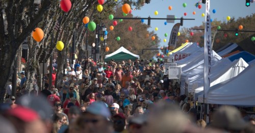 A bustling street festival filled with crowds, colorful balloons, and vendor tents under a clear blue sky.