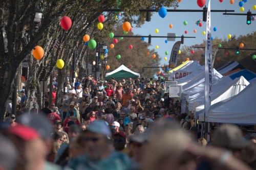 A bustling street fair filled with crowds, colorful balloons, and vendor tents under a clear blue sky.