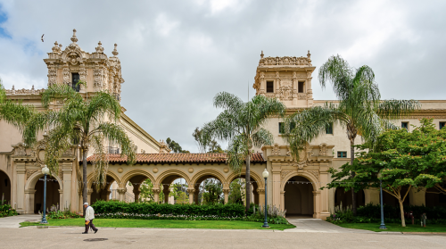 A historic building with ornate architecture, palm trees, and a cloudy sky in a landscaped park setting.