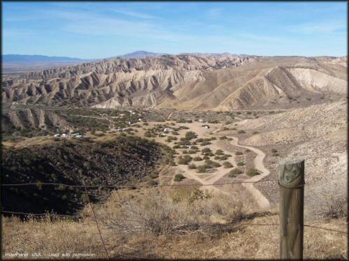 A panoramic view of rugged mountains and valleys under a clear blue sky, with a winding dirt road and sparse vegetation.