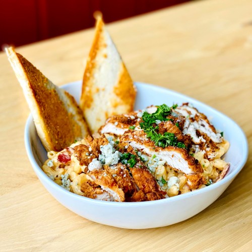 A bowl of creamy macaroni and cheese topped with crispy chicken, herbs, and two slices of toasted bread.