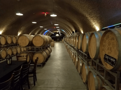 A dimly lit wine cellar with wooden barrels lining the walls and a table set for tasting in the foreground.