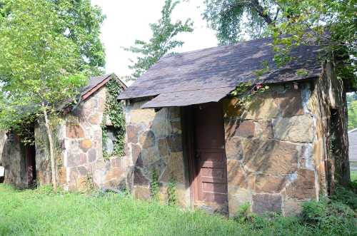 Two small stone buildings with wooden doors, surrounded by greenery and trees.