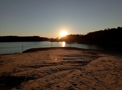 Sunset over a calm lake, with sandy shore and trees silhouetted against the colorful sky.