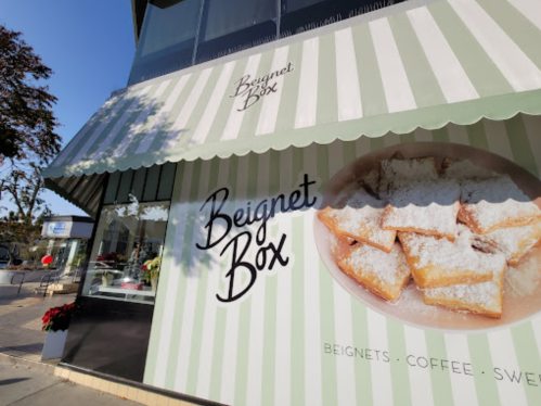 A storefront with a striped awning featuring "Beignet Box" and an image of powdered sugar beignets.