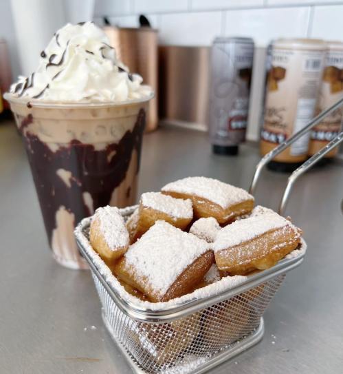 A dessert scene featuring a chocolate milkshake topped with whipped cream and a basket of powdered sugar-dusted pastries.