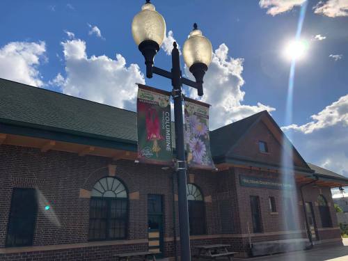 Banners hanging from a lamp post in front of a brick building under a bright sky with clouds.