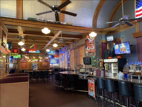 Interior of a bar with wooden beams, a long counter, stools, neon signs, and a TV displaying news.