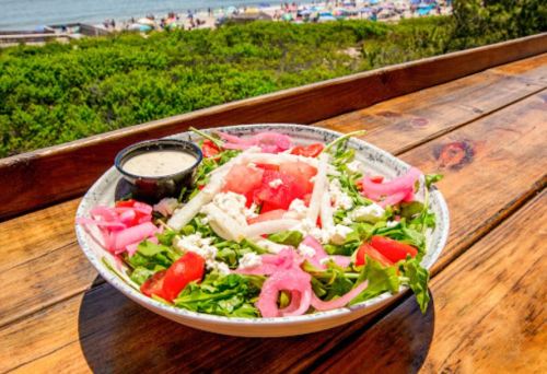 A vibrant salad with greens, tomatoes, watermelon, cheese, and pickled onions, served with dressing, overlooking a beach.