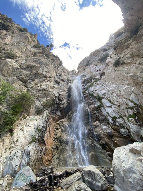 A tall waterfall cascades down rocky cliffs under a bright blue sky with scattered clouds.