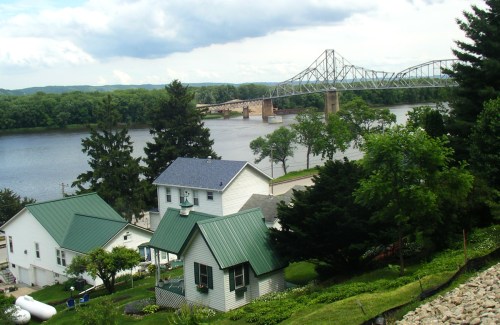 A scenic view of a river with a bridge, surrounded by green trees and houses with metal roofs.