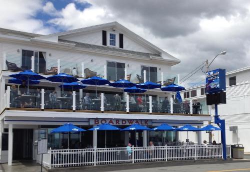 A seaside restaurant with blue umbrellas on the patio, people dining, and a white building under a cloudy sky.