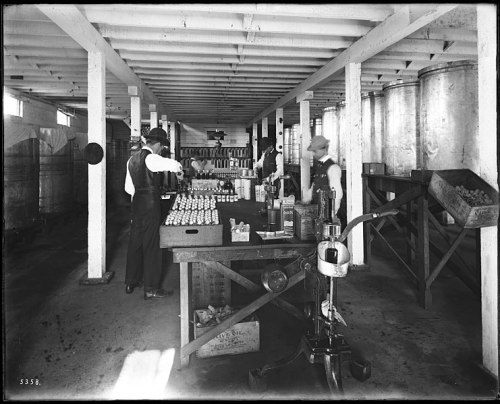 Workers in a vintage factory setting, surrounded by shelves and large containers, engaged in production activities.