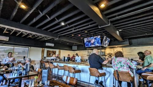 A lively bar scene with patrons seated at the counter and tables, watching a TV screen in a modern, rustic setting.