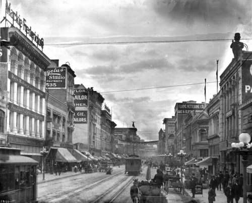 Historic street scene with trams, horse-drawn carriages, and storefronts, set against a cloudy sky.