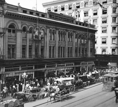 Historic street scene with horse-drawn carriages and a bustling crowd in front of a grand building.