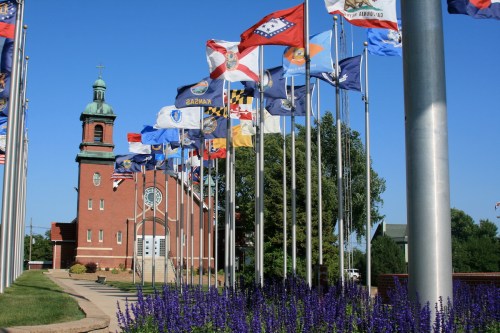 A brick church with a green dome, surrounded by flags and purple flowers under a clear blue sky.