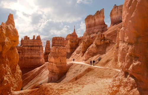 Hikers traverse a winding path through the orange rock formations of Bryce Canyon under a partly cloudy sky.