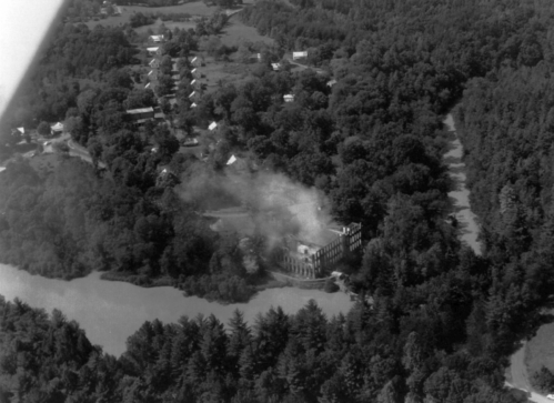 Aerial view of a forested area with a partially obscured building near a lake, surrounded by trees and small structures.