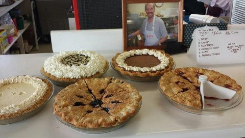 A display of five pies on a counter, with a smiling man in a frame behind them. Pies include cream, chocolate, and fruit varieties.