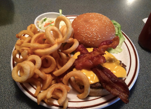A plate with a cheeseburger topped with bacon, lettuce, and a side of curly fries and coleslaw.