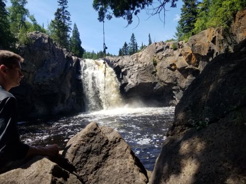 A waterfall cascades over rocky cliffs into a dark pool, surrounded by trees under a clear blue sky. A person sits nearby.