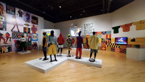 A vibrant exhibition featuring mannequins in colorful outfits, with walls displaying art and multimedia elements.
