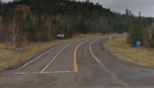 A winding road surrounded by trees and grass, with road signs indicating directions and a cloudy sky above.