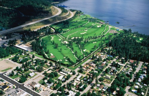 Aerial view of a lush green golf course beside a lake, with a highway and residential area nearby.