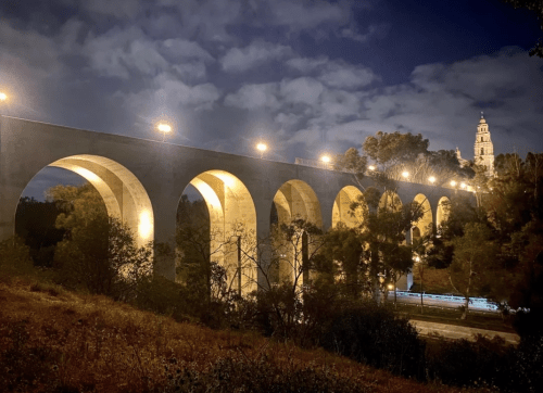 A lit stone aqueduct at night, surrounded by trees and a cloudy sky, with a tower visible in the background.
